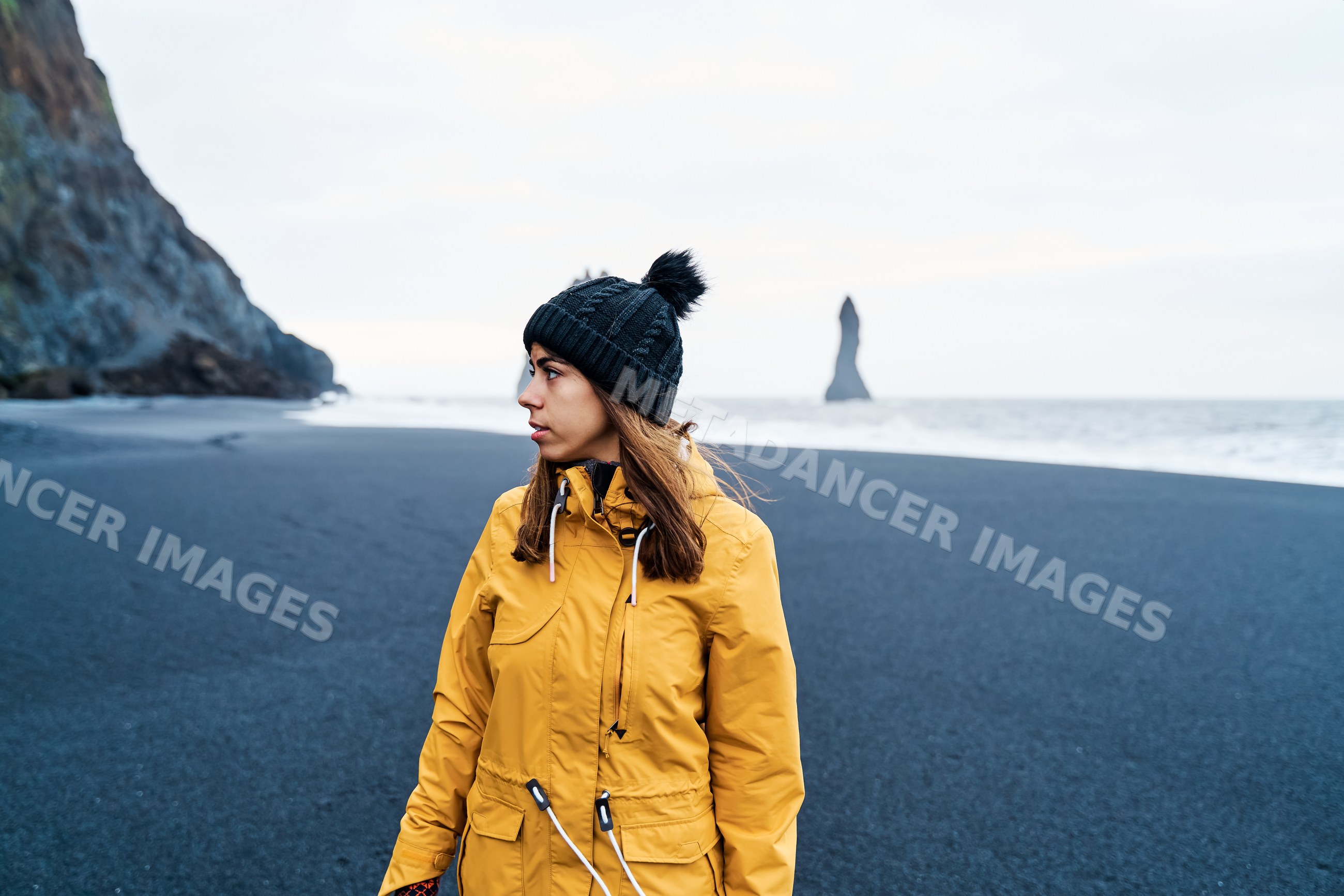 A white woman in winter clothing stands on a black sand beach