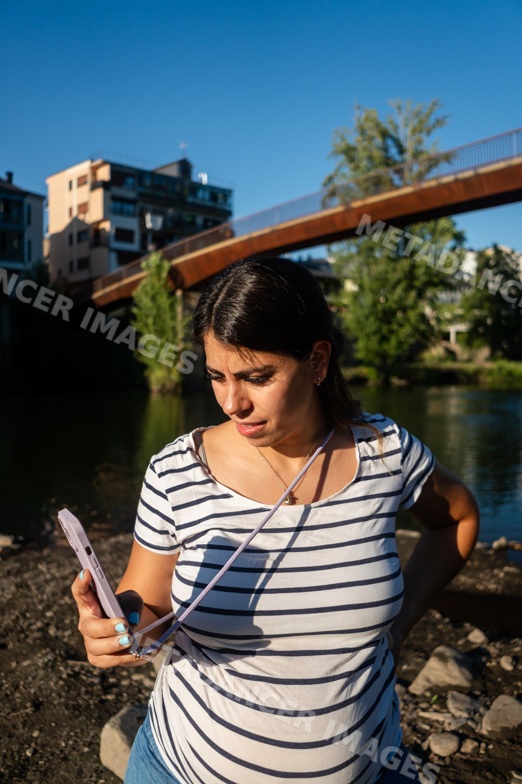 Woman uses phone outdoors by river with bridge and buildings