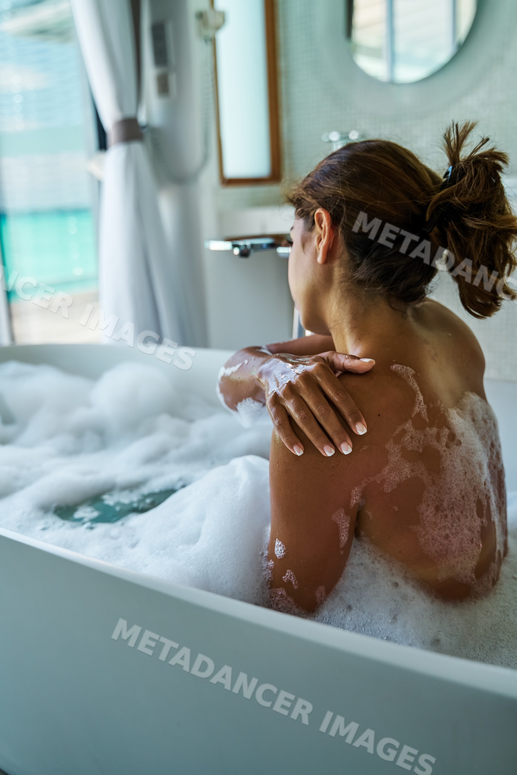 A white woman enjoys a relaxing foamy bath in a modern bathroom