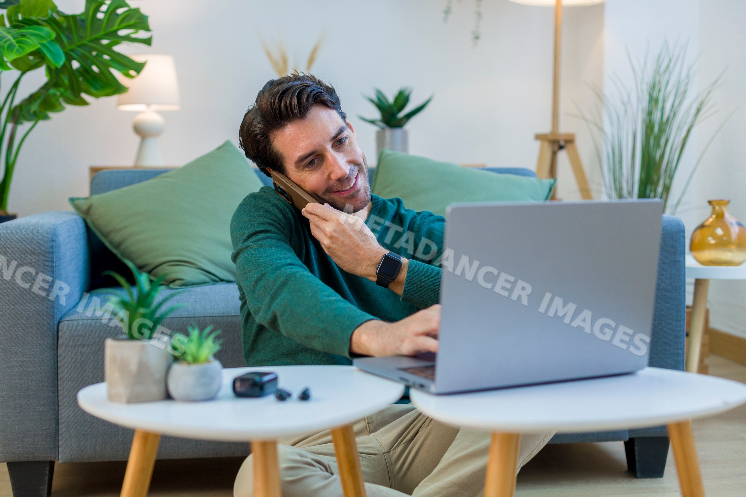White man multitasking with laptop and phone in living room at home