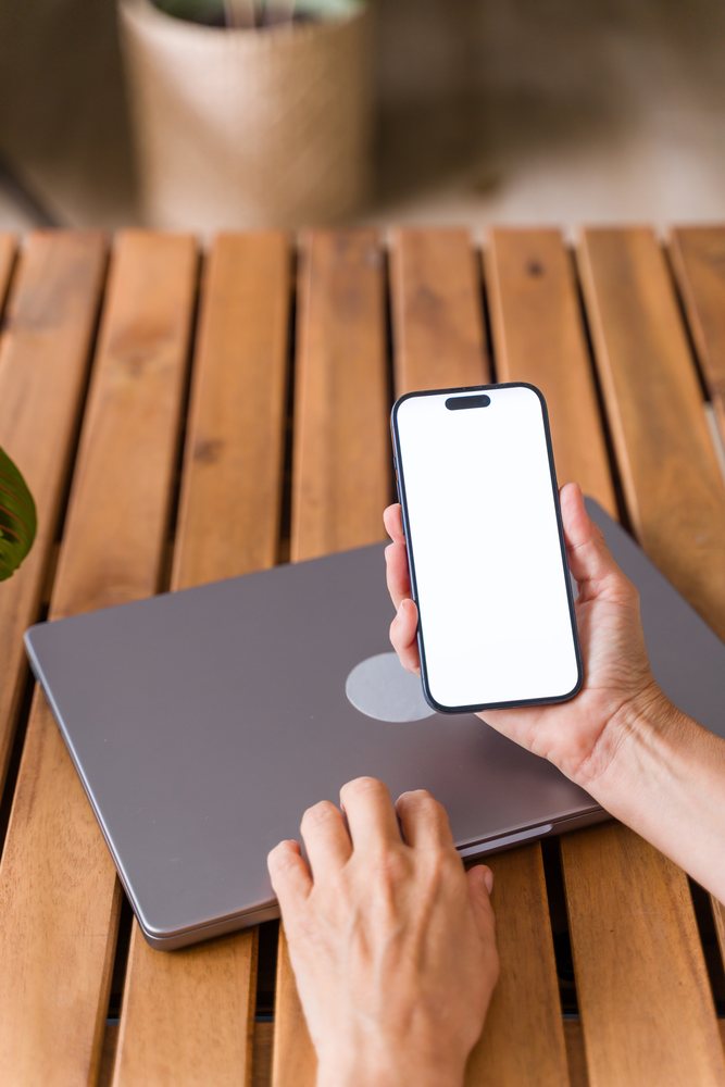 Hand holding smartphone with blank screen above closed laptop