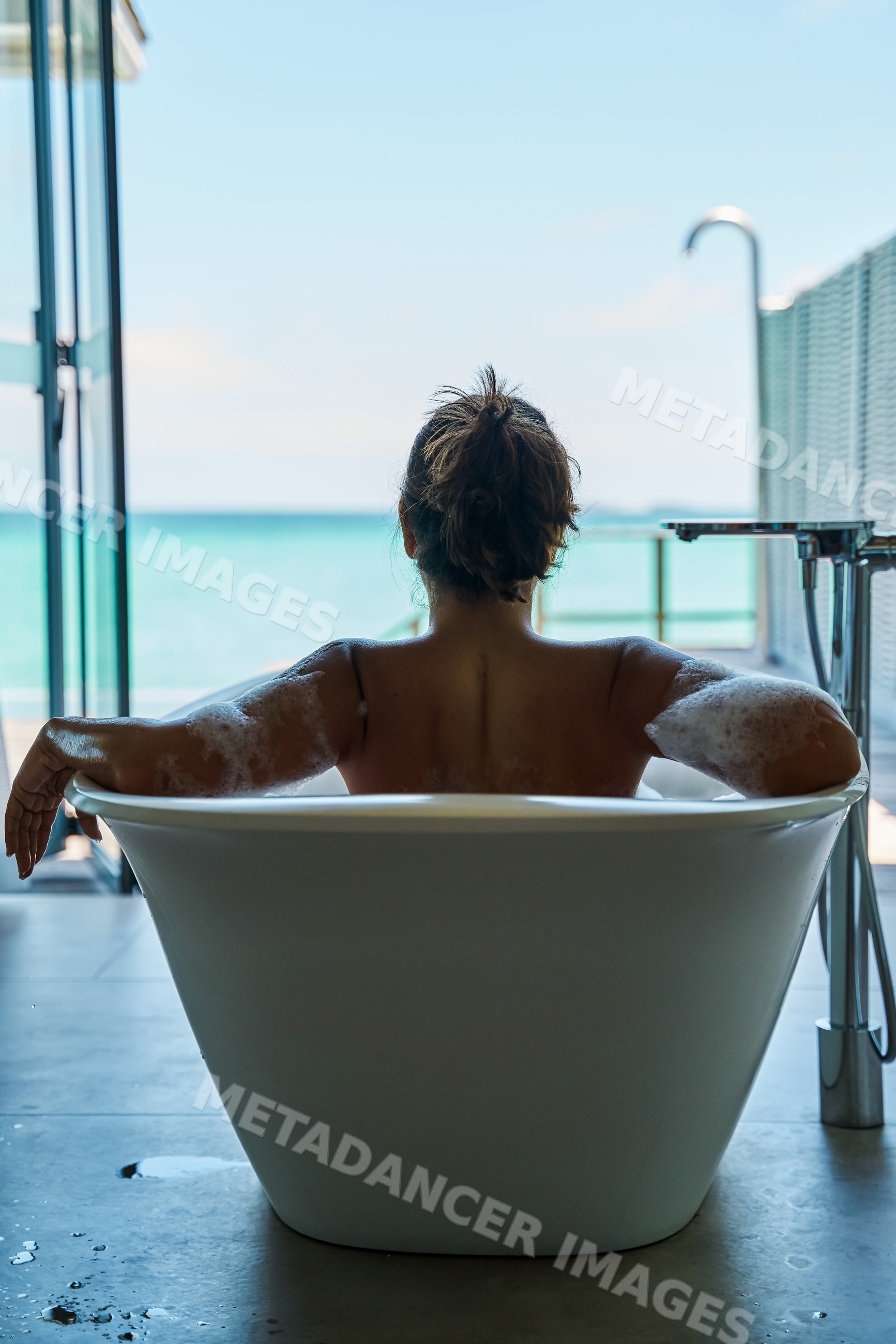 A white woman enjoys a bubble bath in a modern tub with an ocean view