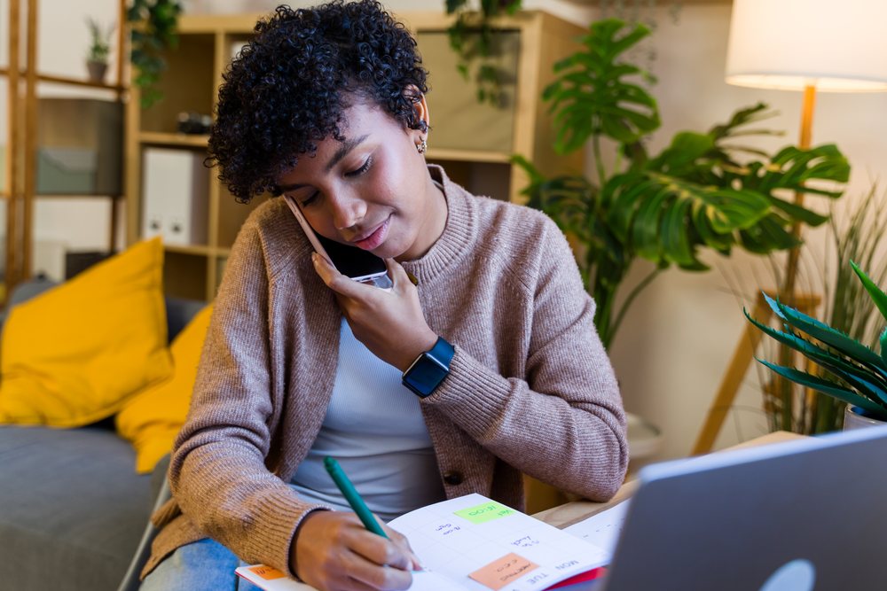 African American woman writing in calendar on phone call at home desk