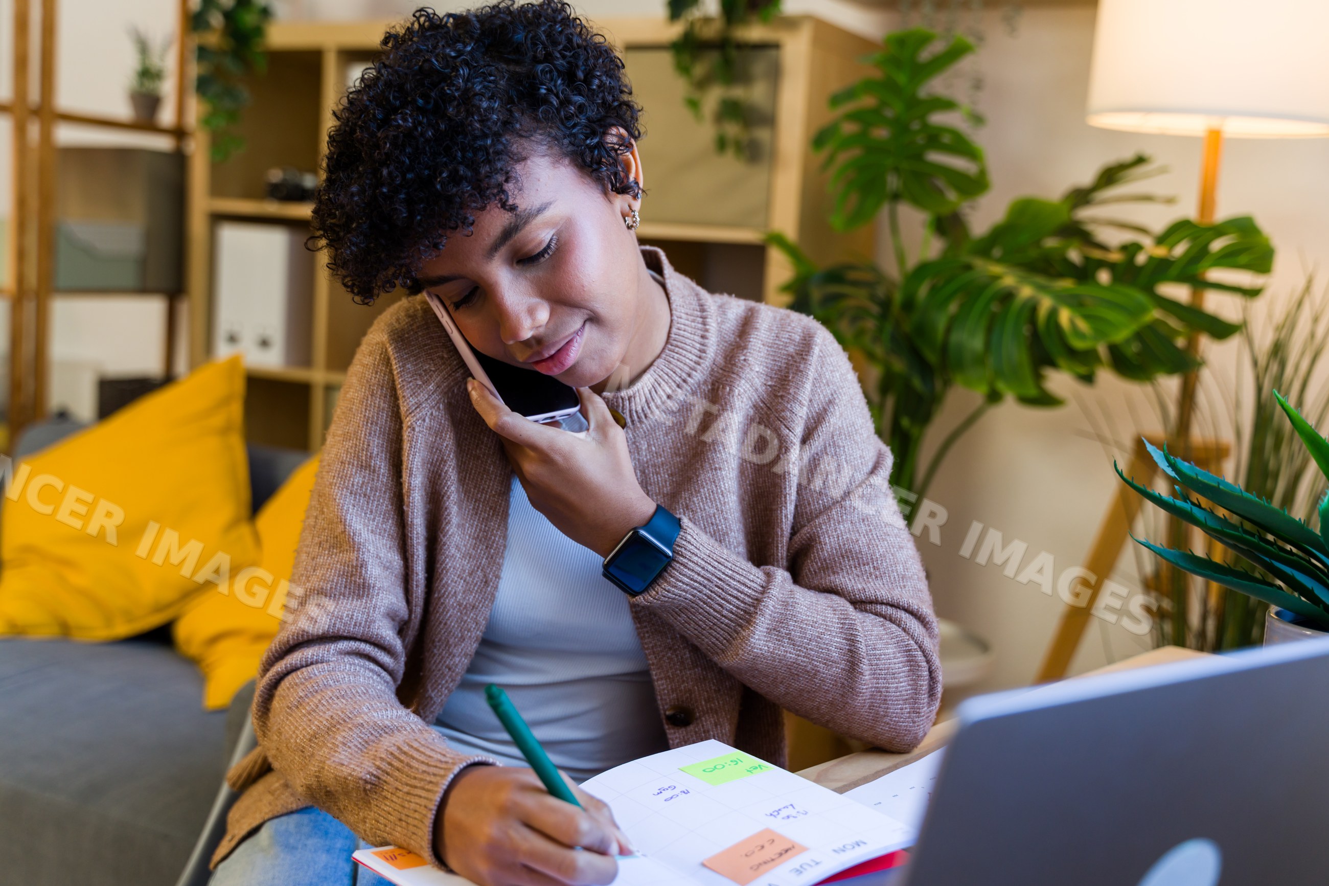 African American woman writing in calendar on phone call at home desk
