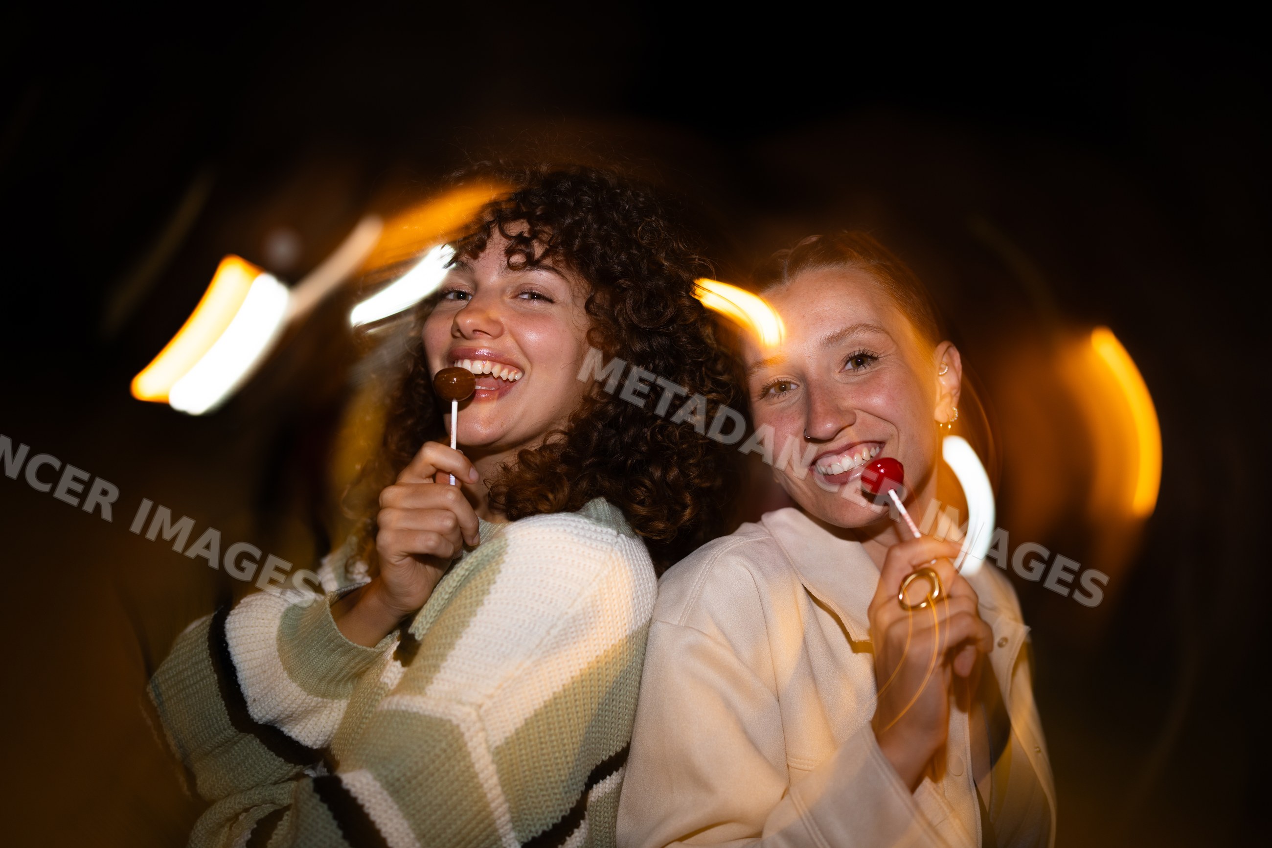 Two white women laughing with lollipops and light trails at night