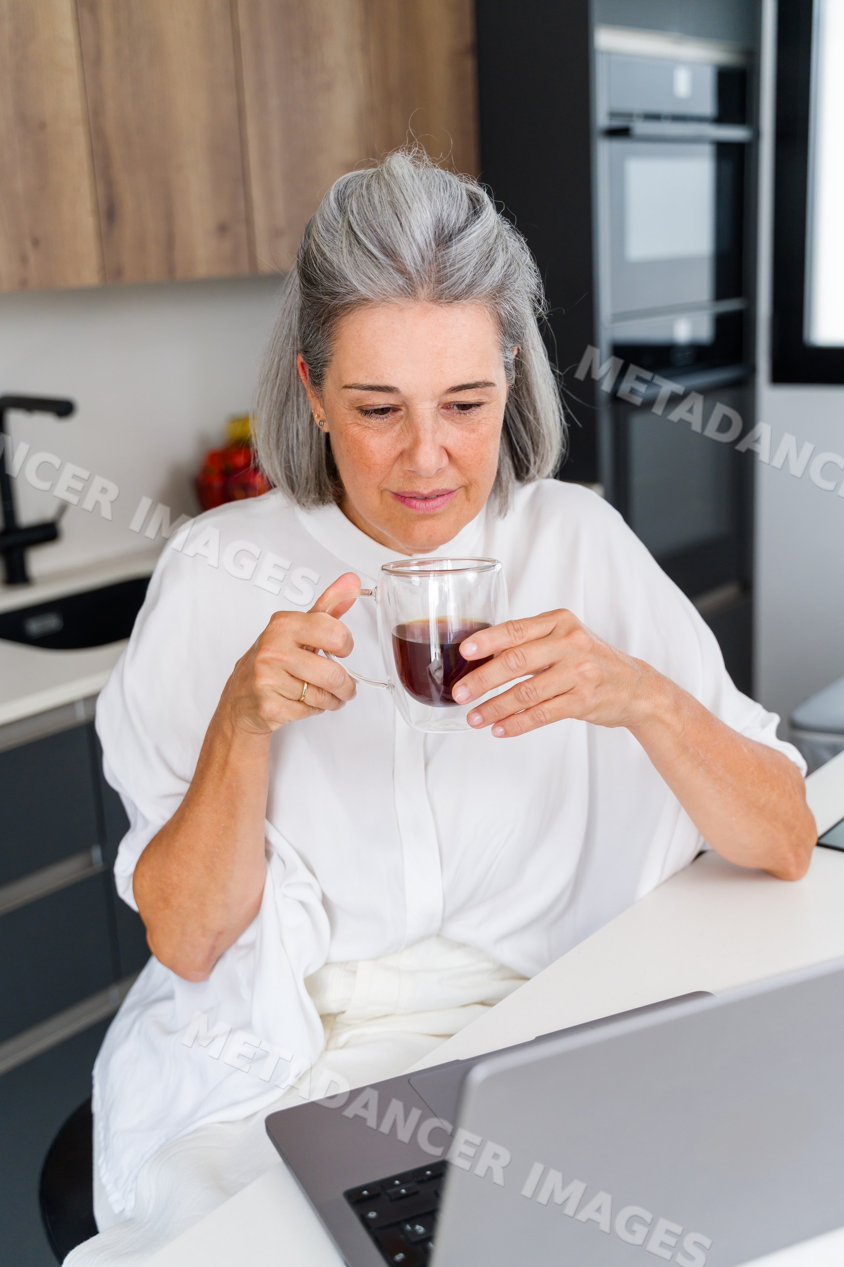 Mature white woman drinking coffee using laptop in modern kitchen