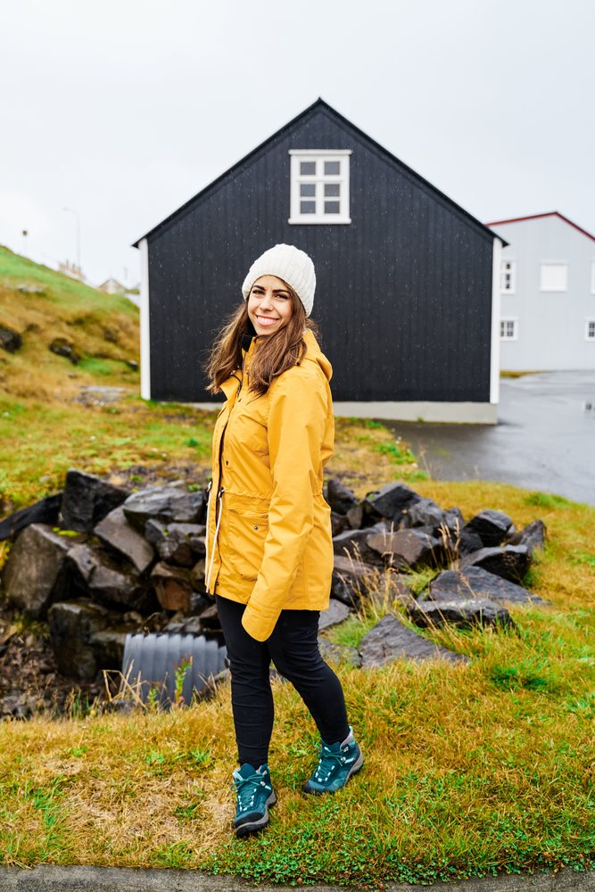 A white woman wearing a yellow jacket walks outside by a black house